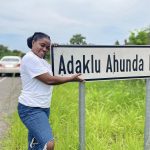 Celestine Agropah smiling proudly next to the Adaklu Ahunda road sign in the Volta Region, marking the community where Happy Daffodils is building its first computer lab.