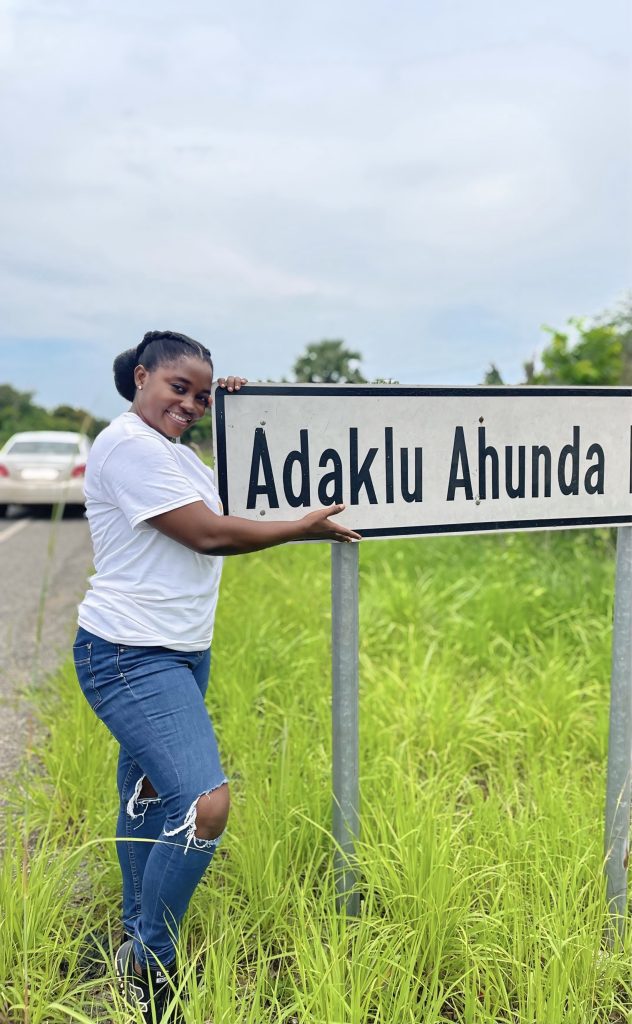 Celestine Agropah smiling proudly next to the Adaklu Ahunda road sign in the Volta Region, marking the community where Happy Daffodils is building its first computer lab.